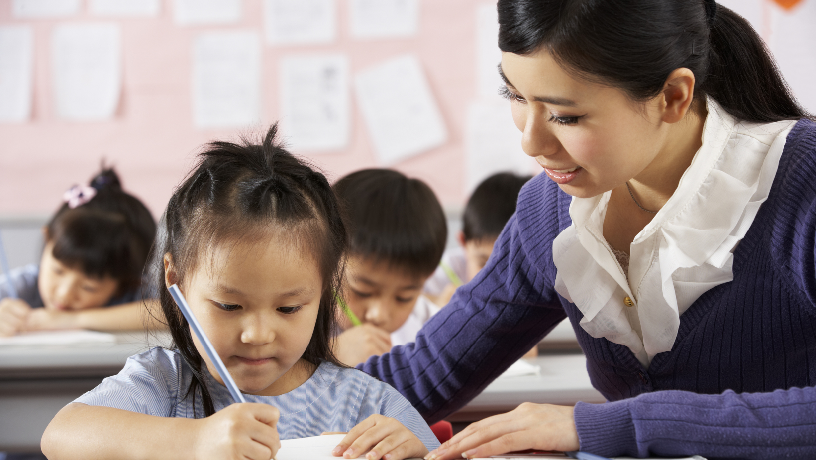 Child writing on paper with teacher looking over shoulder.