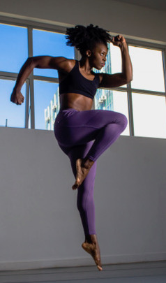 J'nae dressed in a black sports bra and purple tights, balanced on leg and her knee up with the other 