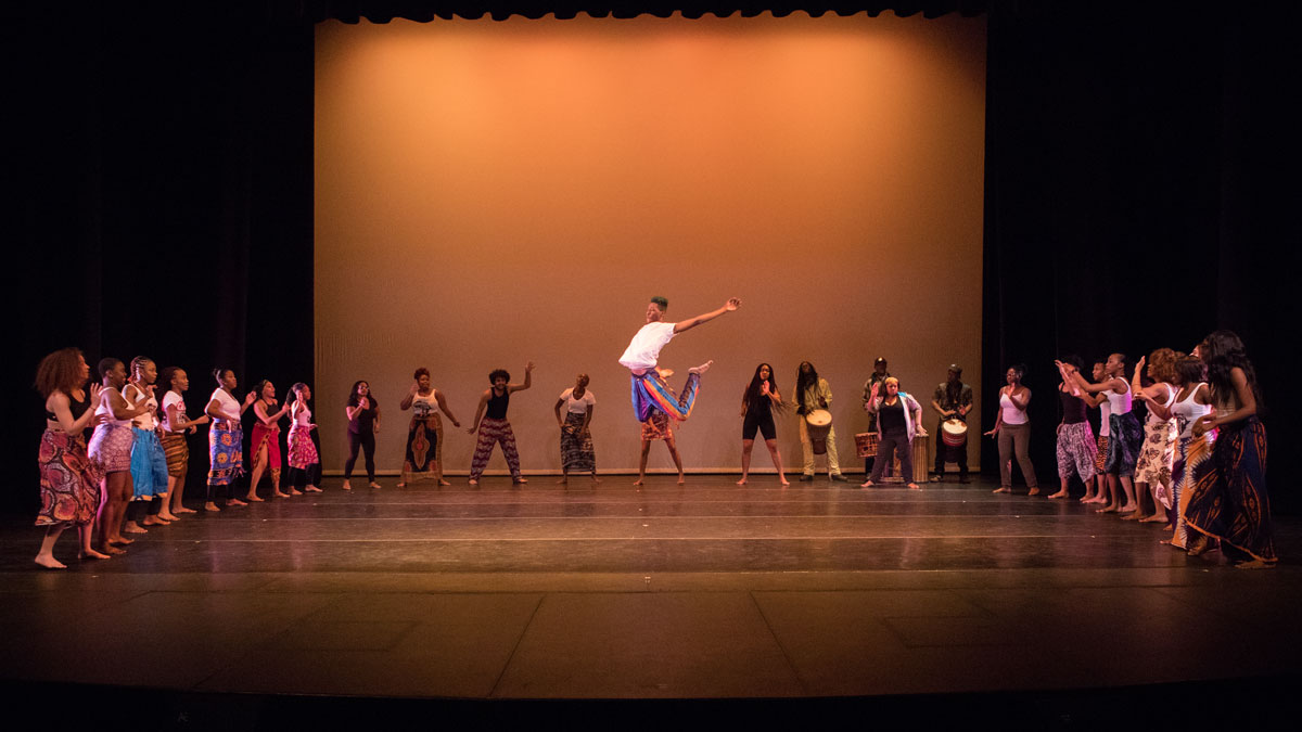 Dancer surround a soloist in a semi-circle formation. The soloist is mid-air in a C jump.