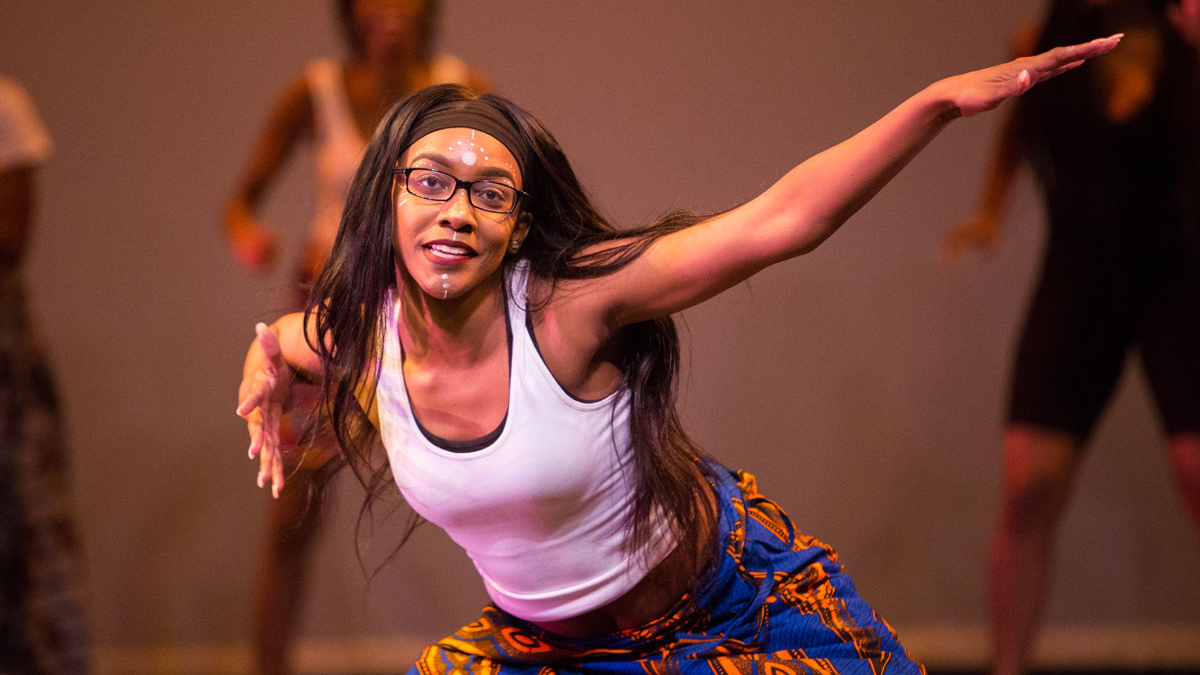 A woman pitched forward with arms extended in the foreground. Dancers are standing behind her in the background.